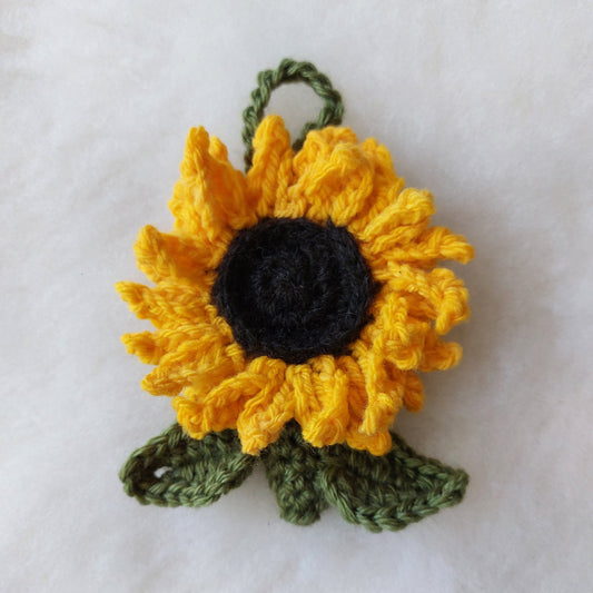 A handmade crocheted sunflower decoration, featuring bright yellow petals, a dark black center, and green leaves, shown on a white textured fabric background.